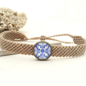 Könnte beinhalten: Ein geflochtenes Armband in Beige mit einem blauen und weißen Fliesen-Anhänger. Der Anhänger ist in einem bronzefarbenen Rahmen gefasst.