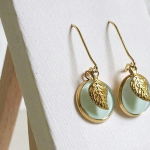 Könnte beinhalten: Ein Paar goldene Ohrringe mit einem hellblauen Glaskreis und einem goldenen Blattanhänger.