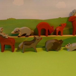 Könnte beinhalten: Ein Set aus Holztierfiguren, darunter ein Reh, ein Waschbär, ein Dachs, ein Wolf, ein Eber und zwei weitere Rehe. Die Figuren sind in Braun-, Grau- und Weißtönen bemalt.
