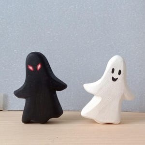 Könnte beinhalten: Zwei Holzfiguren in Form von Geistern. Eine ist schwarz mit roten Augen, die andere ist weiß mit einem lächelnden Gesicht. Die Figuren haben ausgestreckte Arme und stehen auf einer Holzoberfläche, mit einem hellgrauen Hintergrund.