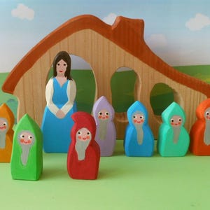 Könnte beinhalten: Ein Holzspielset mit einem Haus mit einer blau gekleideten Frauenfigur und sieben bunten Figuren mit weißen Bärten und Kapuzenmänteln. Die Figuren sind in einer Reihe vor dem Haus angeordnet.