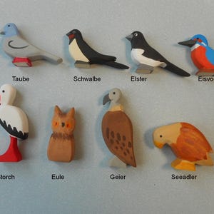Könnte beinhalten: Ein Set aus neun Holzvogel-Figuren, die in verschiedenen Farben bemalt sind, darunter Blau, Rot, Weiß, Schwarz und Braun. Die Figuren sind mit ihren deutschen Namen beschriftet: Taube, Schwalbe, Elster, Eisvogel, Storch, Eule, Geier, Seeadler.