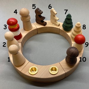 Könnte beinhalten: Holz-Adventskalender mit 10 nummerierten Figuren, darunter ein Leuchtturm, ein Baum, ein Bär und ein Pilz. Die Figuren sind in einem Kreis um zwei Messing-Kerzenhalter angeordnet.
