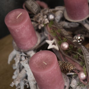 May include: A festive Advent wreath with four mauve pillar candles. The wreath is decorated with white branches, pinecones, ornaments, and a star. Ribbons and greenery add to the holiday theme.