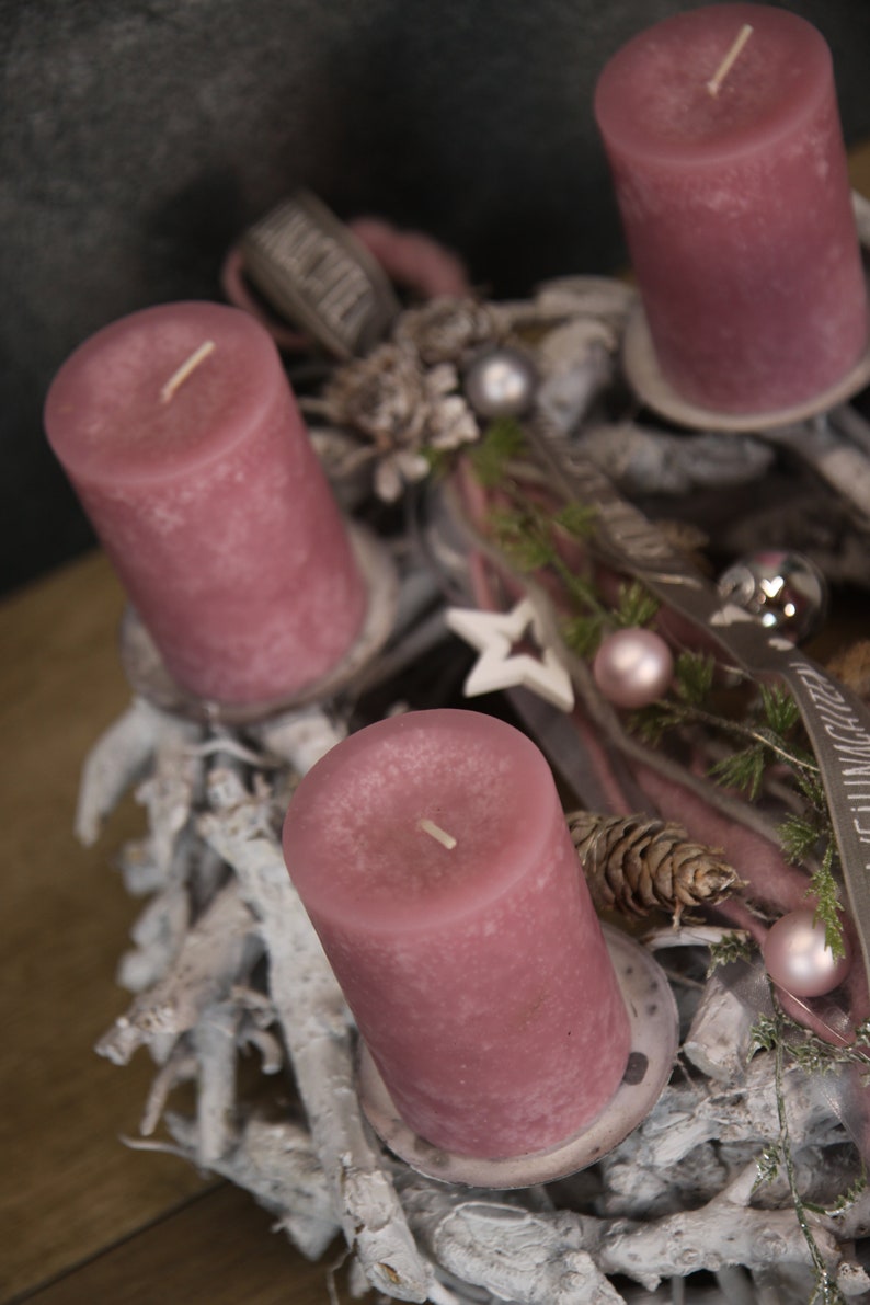 May include: A festive Advent wreath with four mauve pillar candles. The wreath is decorated with white branches, pinecones, ornaments, and a star. Ribbons and greenery add to the holiday theme.