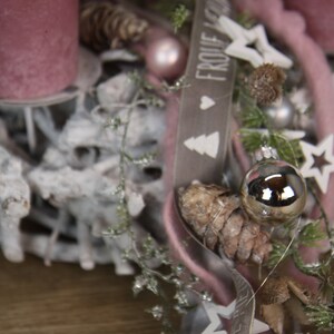 May include: A close-up of a Christmas wreath with a rustic, natural aesthetic. The wreath features a base of white-painted branches, adorned with pink candles, ornaments, pinecones, and a ribbon with the text "FROHE WEIHNACHTEN". Decorative stars and greenery complete the festive design.