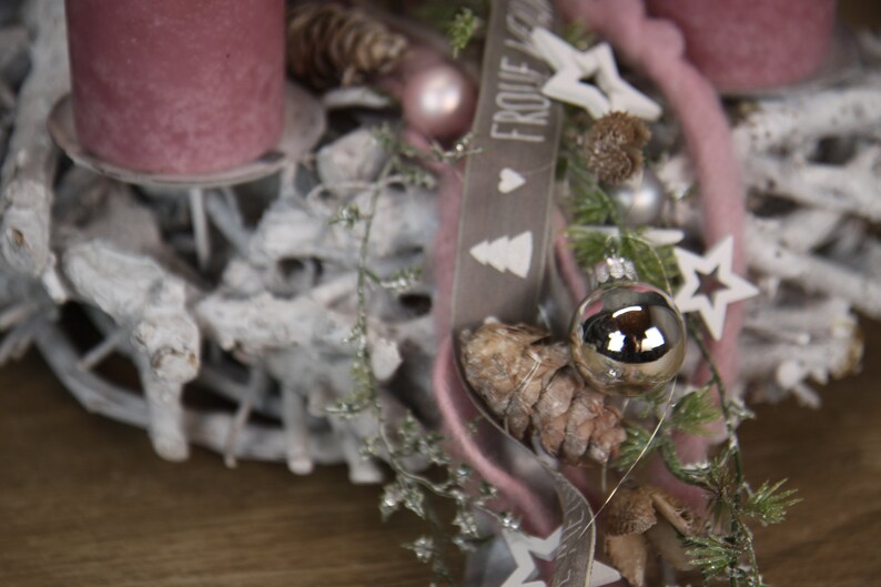 May include: A close-up of a Christmas wreath with a rustic, natural aesthetic. The wreath features a base of white-painted branches, adorned with pink candles, ornaments, pinecones, and a ribbon with the text "FROHE WEIHNACHTEN". Decorative stars and greenery complete the festive design.