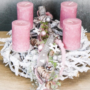 May include: A festive Christmas wreath with four pink pillar candles. The wreath is made of white-painted branches and decorated with silver ornaments, pinecones, and a ribbon with text. A seasonal centerpiece for the holidays.