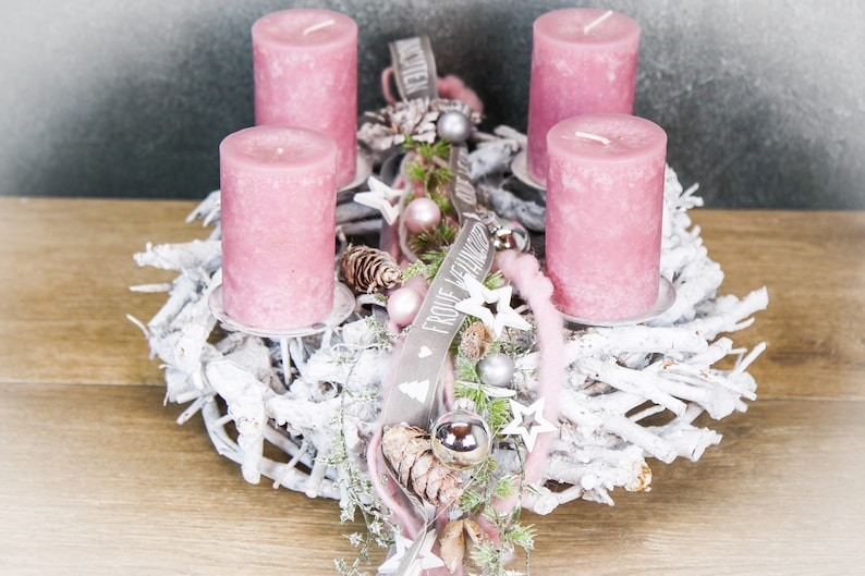 May include: A festive Christmas wreath with four pink pillar candles. The wreath is made of white-painted branches and decorated with silver ornaments, pinecones, and a ribbon with text. A seasonal centerpiece for the holidays.