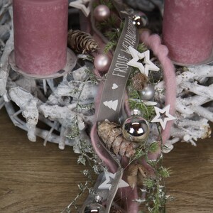 May include: A festive Christmas wreath with two mauve candles. The wreath is made of white-painted branches and decorated with silver ornaments, stars, pinecones, and a ribbon with the text "FROHE WEIHNACHTEN".