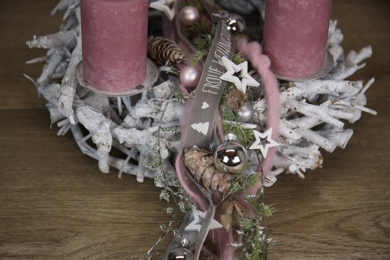 May include: A festive Christmas wreath with two mauve candles. The wreath is made of white-painted branches and decorated with silver ornaments, stars, pinecones, and a ribbon with the text "FROHE WEIHNACHTEN".