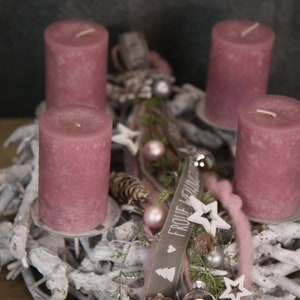 May include: A festive Advent wreath with four mauve candles. The wreath is decorated with white branches, pinecones, stars, and ornaments. A small wooden sign with text is also included. The overall theme is holiday decor.