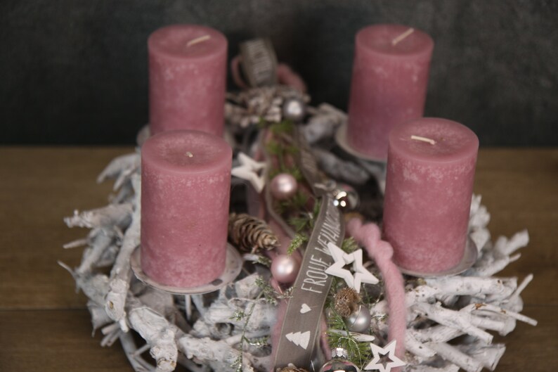 May include: A festive Advent wreath with four mauve candles. The wreath is decorated with white branches, pinecones, stars, and ornaments. A small wooden sign with text is also included. The overall theme is holiday decor.