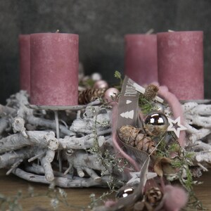 May include: A Christmas wreath with four mauve candles. The wreath is made of white branches and decorated with pinecones, silver ornaments, and a ribbon with text. A star and other decorative elements complete the festive arrangement.