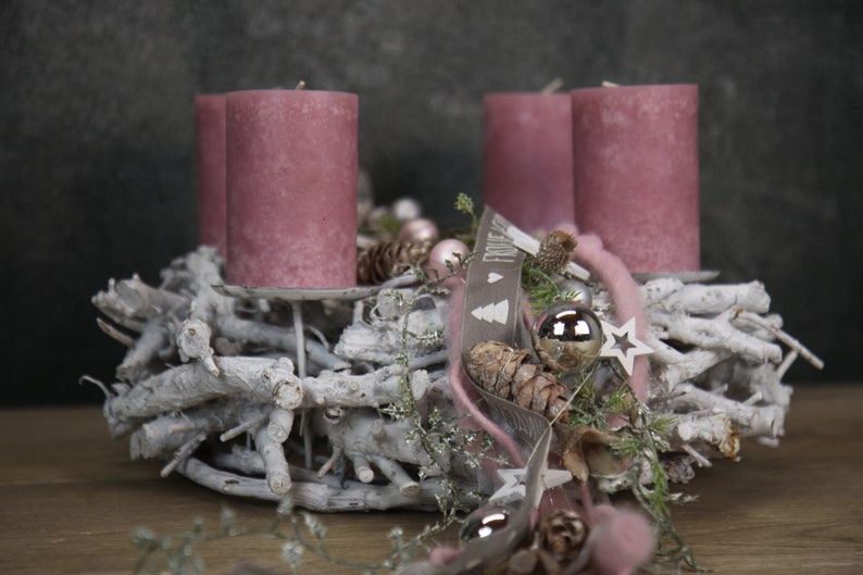 May include: A Christmas wreath with four mauve candles. The wreath is made of white branches and decorated with pinecones, silver ornaments, and a ribbon with text. A star and other decorative elements complete the festive arrangement.