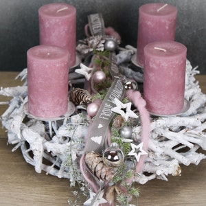 May include: A festive Advent wreath with four mauve candles. The wreath is made of white-painted branches and decorated with silver ornaments, stars, and a ribbon that says "Frohe Weihnachten".
