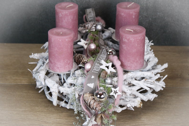 May include: A festive Advent wreath with four mauve candles. The wreath is made of white-painted branches and decorated with silver ornaments, stars, and a ribbon that says "Frohe Weihnachten".