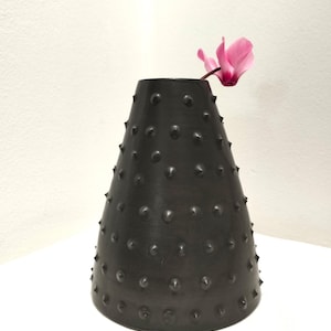 Könnte beinhalten: Eine schwarze Keramikvase mit einer strukturierten, mit Nieten besetzten Oberfläche. Eine einzelne rosa Blume steht in der Vase.