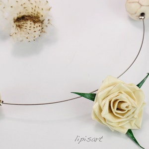 May include: A delicate necklace featuring a cream-colored rose and three off-white, patterned beads. The necklace has a thin, silver-colored wire and green leaves. The word "lipisart" is written in cursive.