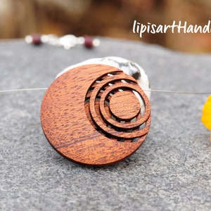 Könnte beinhalten: Eine Holz-Anhänger-Halskette mit einem kreisförmigen Design. Der Anhänger besteht aus rotbraunem Holz und weist konzentrische Kreise auf. Die Halskette wird auf einer grauen Oberfläche mit einer gelben Blume und dem Text "lipisartHandmade" präsentiert.