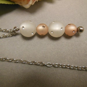 Puede incluir: Un collar plateado con una serie de cuentas blancas esmeriladas y color melocotón, cada una adornada con pequeños cristales brillantes. Las cuentas están ensartadas y conectadas a una delicada cadena.