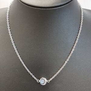 Könnte beinhalten: Eine zarte Silberkette mit einem kleinen, runden Anhänger mit einem hellblauen Stein. Der Anhänger hängt an einer feinen Kette und erzeugt ein schlichtes und elegantes Design. Die Halskette wird auf einer schwarzen Schaufensterpuppe präsentiert.