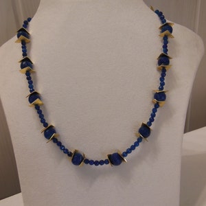 Puede incluir: Un collar de cuentas azules con detalles dorados. El collar presenta grandes cuentas azules redondas intercaladas con cuentas azules más pequeñas y formas geométricas doradas. El collar se exhibe en un maniquí blanco.