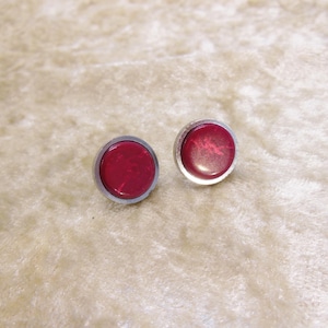 Op de afbeelding: Een paar ronde oorstekers met een zilverkleurige metalen zetting. De oorbellen hebben een dieprood, gemarmerd effect in het midden. De oorbellen worden gepresenteerd op een getextureerd, lichtbeige oppervlak.