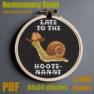 May include: A cross-stitch pattern in a wooden hoop features a snail wearing a cowboy hat. The text "LATE TO THE" and "HOOTENANNY" are stitched above and below the snail. The pattern is 84x84 stitches and uses 11 DMC colors.