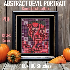 May include: A cross stitch pattern of an abstract devil portrait in red, purple, and black. The pattern is 100x100 stitches and uses 13 DMC colors. The pattern is framed in a black frame and is sitting on a wooden surface.