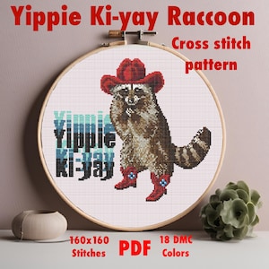 Pode incluir: Padrão de ponto cruz de um guaxinim usando um chapéu de caubói vermelho e botas, com o texto "Yippie Ki-yay" em azul e branco. O padrão é de 160x160 pontos e usa 18 cores DMC.