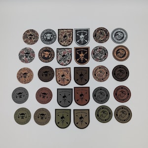 May include: A collection of military-themed patches with various designs. Some patches are circular, featuring skulls and text, while others are shield-shaped with crossed arrows and text. The color palette includes shades of brown, green, and gray.