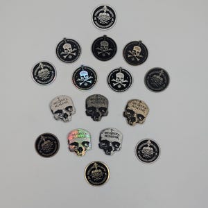 May include: A collection of circular and skull-shaped patches featuring various finishes including metallic, holographic, and matte black. The circular patches display a skull and crossbones with a cross and text. The skull patches feature the text MONAXOS.