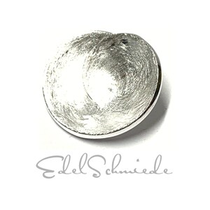 Könnte beinhalten: Ein silberfarbener, kreisförmiger Anhänger mit einer strukturierten, gebürsteten Metalloberfläche. Der Anhänger hat ein leicht konkaves Design und einen glatten, polierten Rand. Der Text "Edel Schmiede" steht darunter.