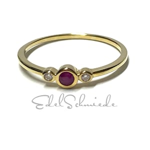 Könnte beinhalten: Ein goldener Ring mit einem zentralen Rubin und zwei kleineren Diamanten. Der Ring hat ein zartes Band und ist vor weißem Hintergrund platziert. Der Text "Edel Schmiede" ist darunter zu sehen.