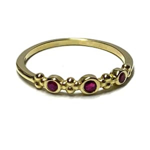 Könnte beinhalten: Ein goldener Ring mit drei kleinen, runden, tiefrosa Rubinen in Zargenfassungen. Das Band ist dünn und poliert, wobei kleine dekorative Goldkugeln zwischen jedem der drei Steine platziert sind, was ein zartes und symmetrisches Design ergibt.
