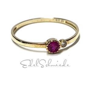 Könnte beinhalten: Ein goldener Ring mit einem leuchtend roten Edelstein und einem kleineren, klaren Stein. Der Ring hat ein schmales Band und ist vor einem weißen Hintergrund platziert. Der Text "Edel Schmiede" ist darunter zu sehen.