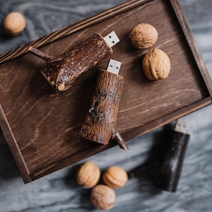 May include: Two wooden USB drives shaped like tree branches with bark detail. The drives are on a wooden surface with walnuts scattered around them.