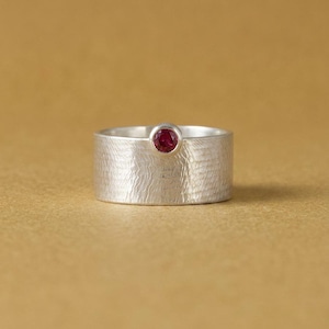 Könnte beinhalten: Ein breiter Silberring mit einer texturierten, gebürsteten Metalloberfläche und einem einzelnen, kleinen, runden, tiefroten Edelstein, der in einer glatten Fassung oben in der Mitte des Rings sitzt. Der Ring hat ein flaches Profil und eine gleichmäßige Breite.
