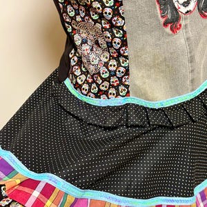 Puede incluir: Un delantal colorido con el tema del Día de Muertos. El delantal tiene una falda de lunares negros, un corpiño con estampado de calaveras y una parte superior de mezclilla. El diseño incluye una calavera de pedrería y una aplicación de calavera de azúcar.