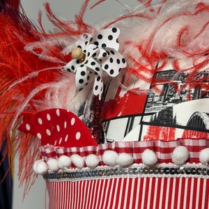 Könnte beinhalten: Festlicher Kopfschmuck mit roten und weißen Federn, einer gepunkteten Schleife und einer rot-weiß gestreiften Basis. Der Kopfschmuck ist mit weißen Pompons und einem rot-weiß-schwarzen Muster verziert.