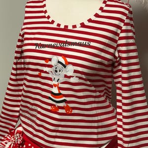 Puede incluir: Una camiseta de manga larga a rayas rojas y blancas con el texto "Karnevalsmaus" y un gráfico de ratón de dibujos animados. El conjunto incluye una falda de lunares rojos y un sombrero a juego, creando un conjunto festivo.