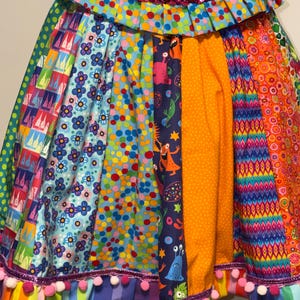 以下が含まれることがあります： デニムのウエストバンドとスパンコールトリムが付いた、カラフルなパッチワークスカート。スカートは、水玉模様、花柄、モンスターのデザインなど、さまざまな模様の生地が特徴で、裾にポンポンが付いています。スカートは活気があり遊び心のある衣服です。