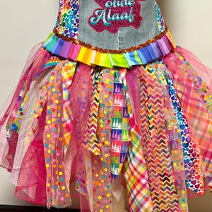 May include: A vibrant tutu dress with a denim bodice embroidered with the text "Niemals ohne Alaaf". The skirt features layers of pink tulle, patterned ribbons, and fabric in various colours and designs, including polka dots, hearts, and zigzags.