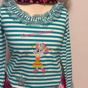Puede incluir: Una camiseta de manga larga a rayas con un dibujo de ratón de dibujos animados y el texto "Karnevalsmaus". La camiseta tiene un cuello con volantes y se combina con una falda patchwork. Se ve un sombrero decorativo con plumas rosas y una etiqueta en forma de corazón.