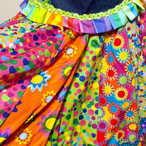Puede incluir: Una falda con una cintura de mezclilla azul oscuro con un borde verde lima, seguida de un vibrante mosaico de telas multicolores. Los patrones incluyen corazones rosas, girasoles naranjas y lunares multicolores, terminada con un borde de pompones azul cerceta.
