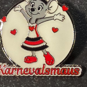 Könnte beinhalten: Anstecknadel mit einer Cartoon-Maus mit grauem Fell, die ein rot-schwarzes Kleid und Schuhe sowie einen passenden Hut trägt. Die Maus ist vor einem weißen Kreis mit roten Herzakzenten und dem Wort "Karnevalsmaus" in Rot platziert.