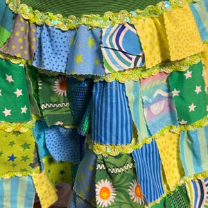 Puede incluir: Una colorida falda patchwork con cinturilla verde y capas de tela en azul, verde y amarillo. La falda presenta varios estampados, incluyendo estrellas, lunares y margaritas, con un ribete decorativo.