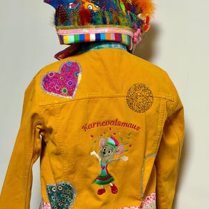 Könnte beinhalten: Senfgelbe Jeansjacke mit bunten Aufnähern und Stickereien. Auf der Rückseite befindet sich eine Cartoon-Maus und der Text "Karnevalsmaus". Die Jacke ist mit einem dekorativen Hut mit Federn und farbigen Stoffbesätzen verziert. Die Jacke hat ein skurriles, festliches Design.