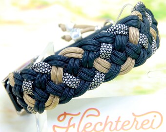 May include: A paracord collar featuring a braided pattern in black, tan, and a reflective silver-flecked black cord. A white tag is attached with the text Flechterei Rund um Hund in orange and gray lettering.
